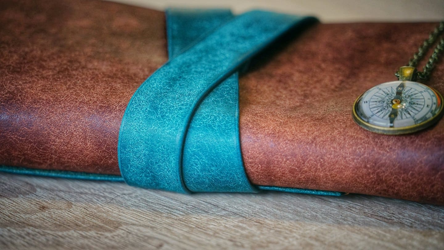 Brown leather journal with teal strap and compass pendant on a wooden table.