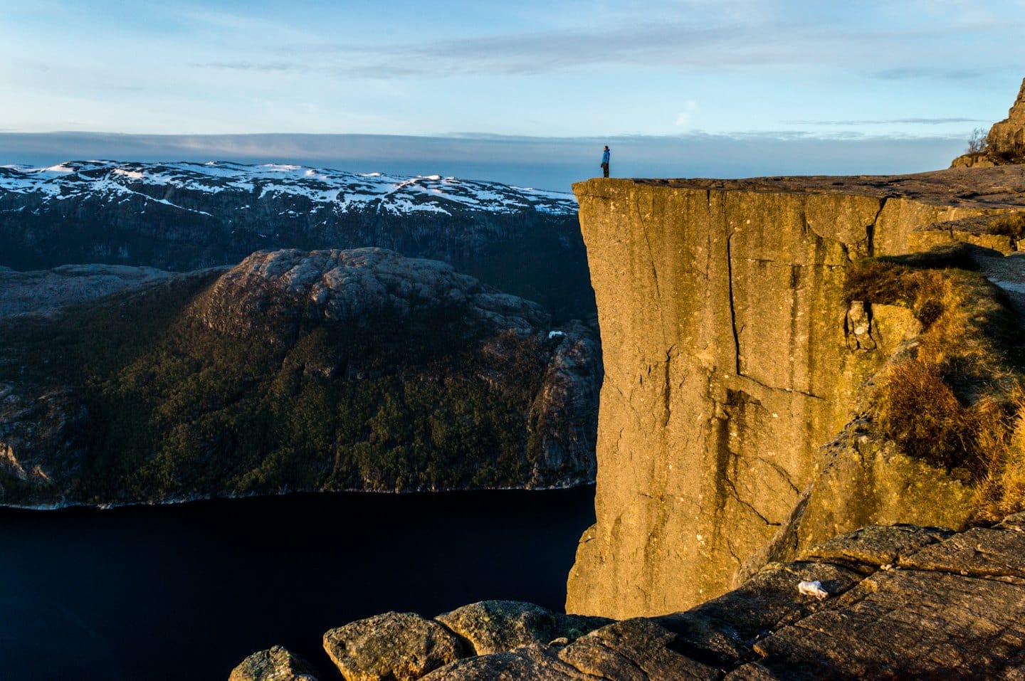 A hiker stands on a cliff edge overlooking a mountain fjord.